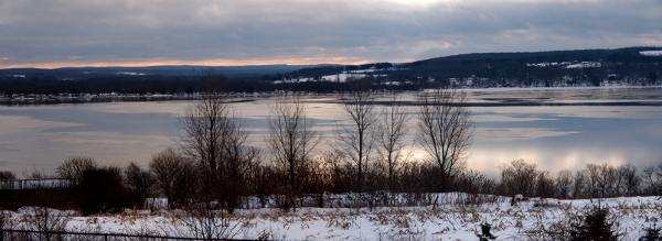 [Several photos stitched together to give a view of the partially frozen lake under a very cloudy sky at sunset with just enough breaks in the clouds to see a partially reflection of them on the water.]